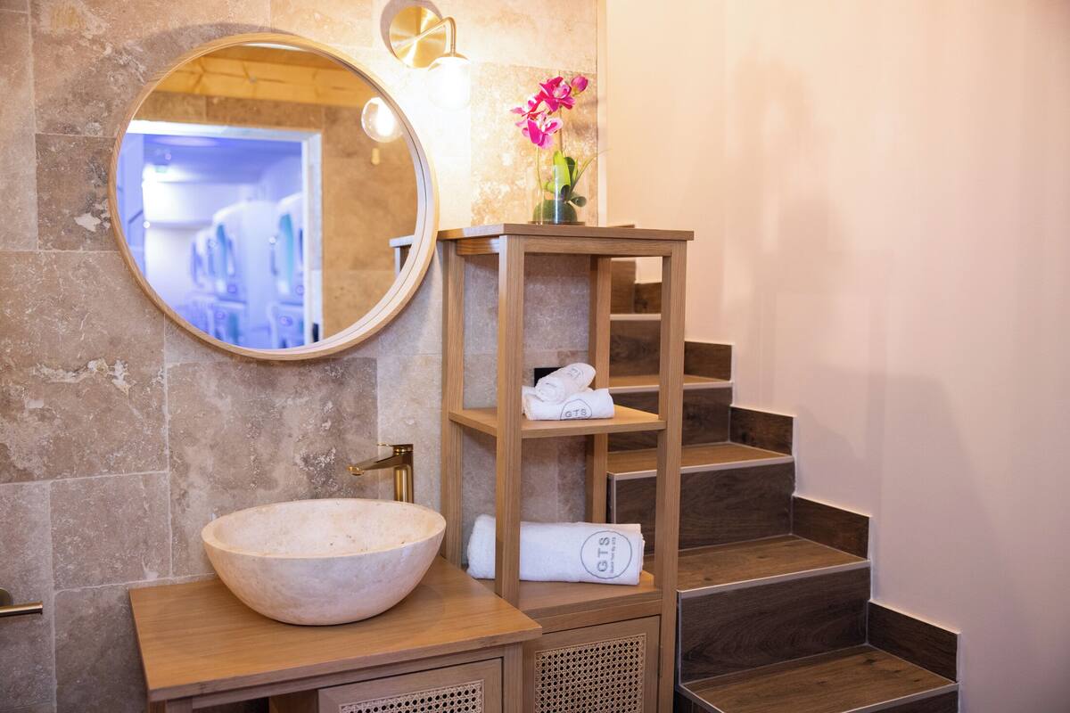 Un baño con un espejo redondo, un tocador de madera con un lavabo grande tipo cuenco y un estante de madera con toallas.