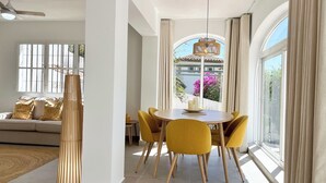 Un coin repas avec une table ronde, des chaises jaunes et une vue sur un bâtiment orné de fleurs roses à l’extérieur.