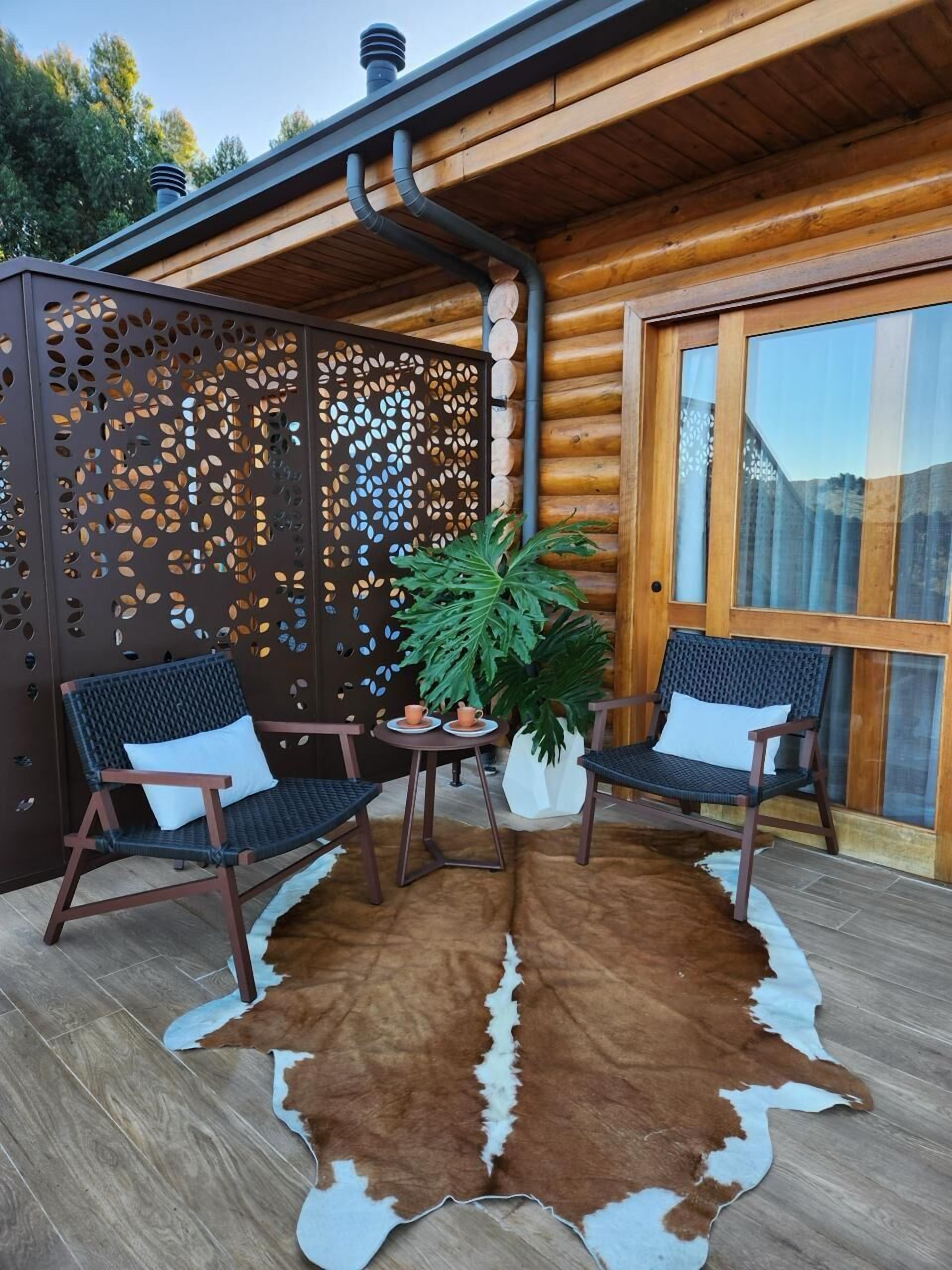 A wooden cabin with a covered patio featuring two wicker chairs, a table, and a potted plant.