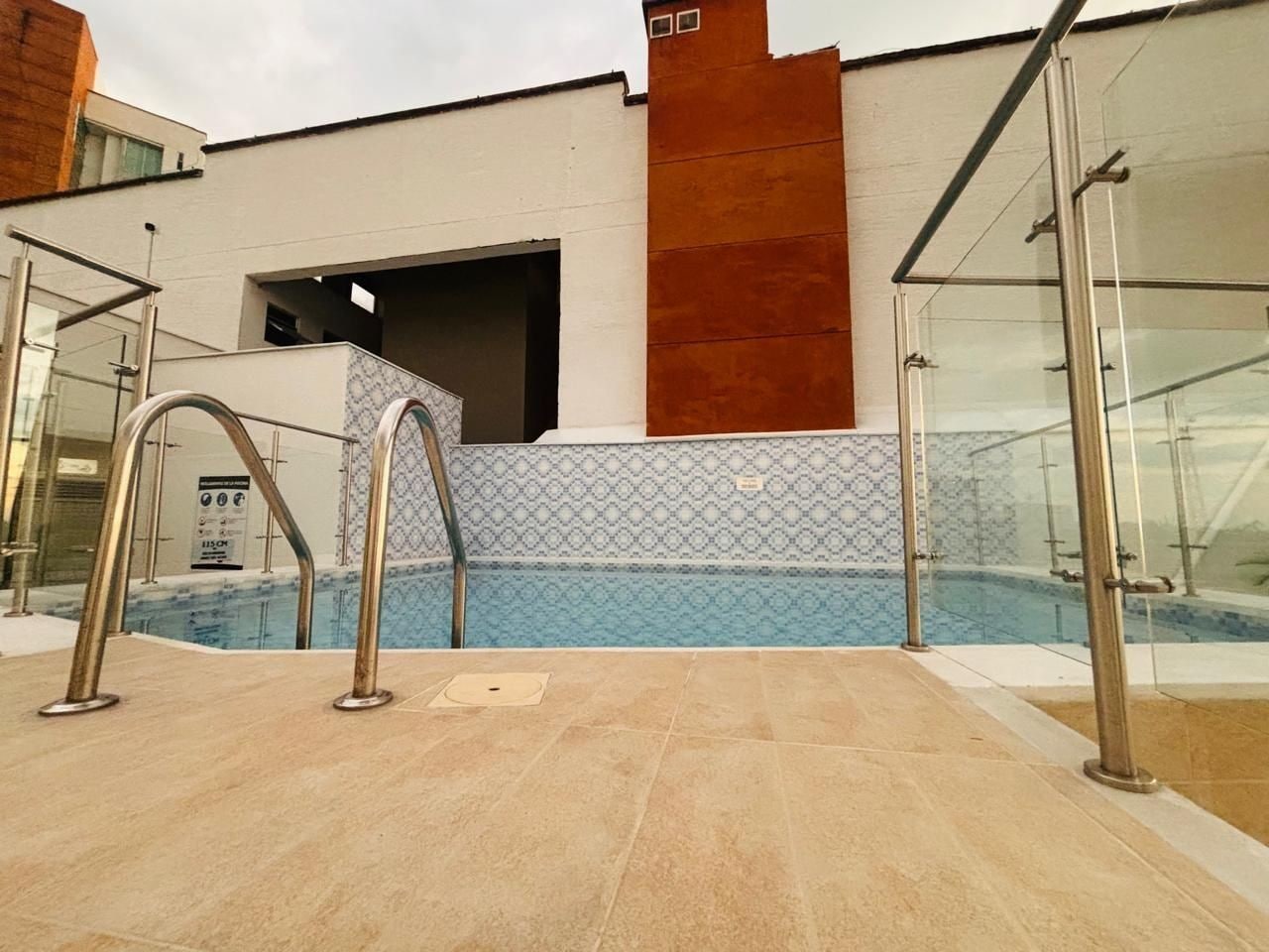 An indoor swimming pool with clear glass railings, a tiled floor, and a modern building facade with a patterned wall.