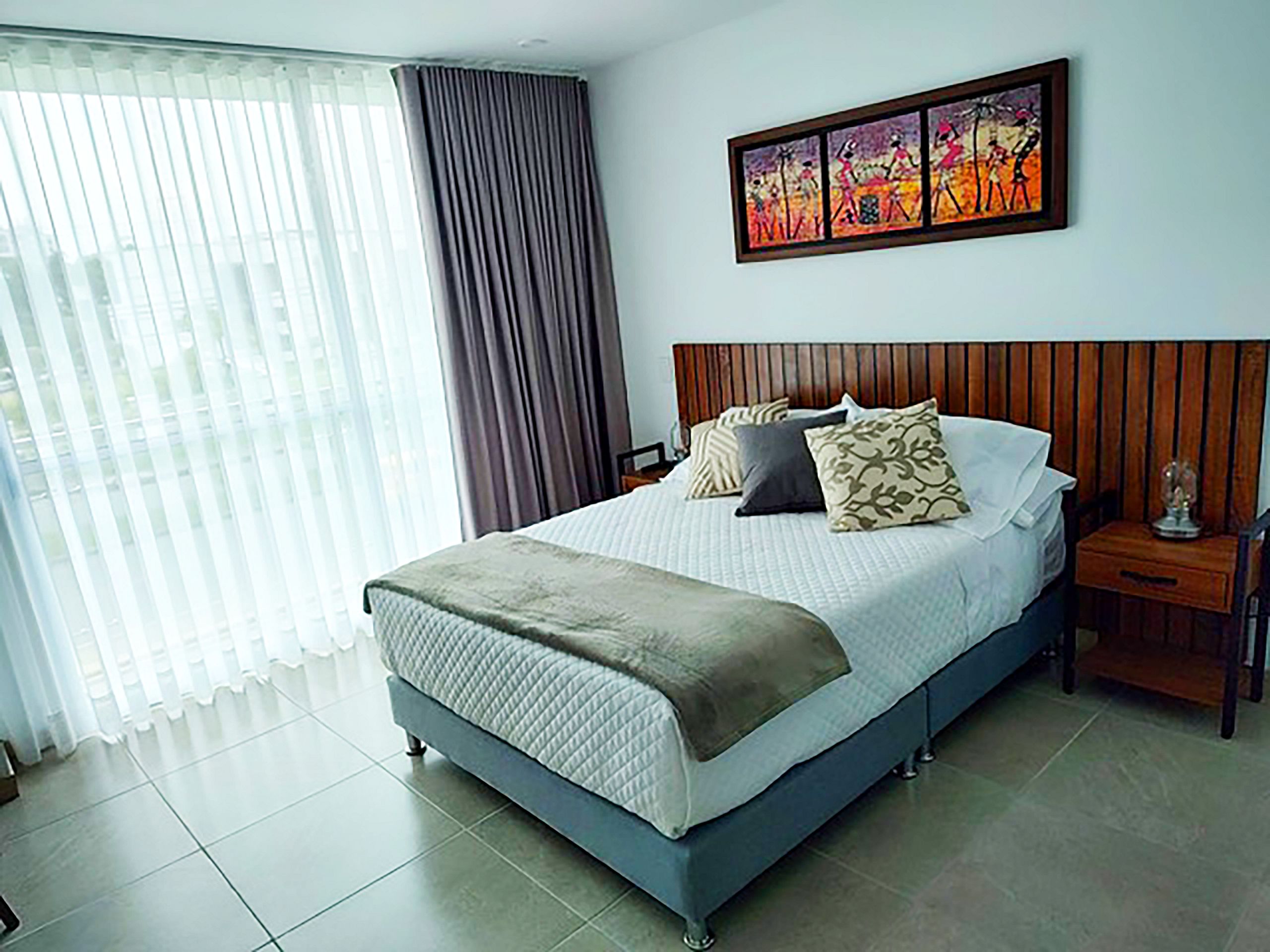 Une chambre d’hôtel avec un grand lit, une tête de lit, une table de chevet, un tableau au mur et une fenêtre avec des rideaux.