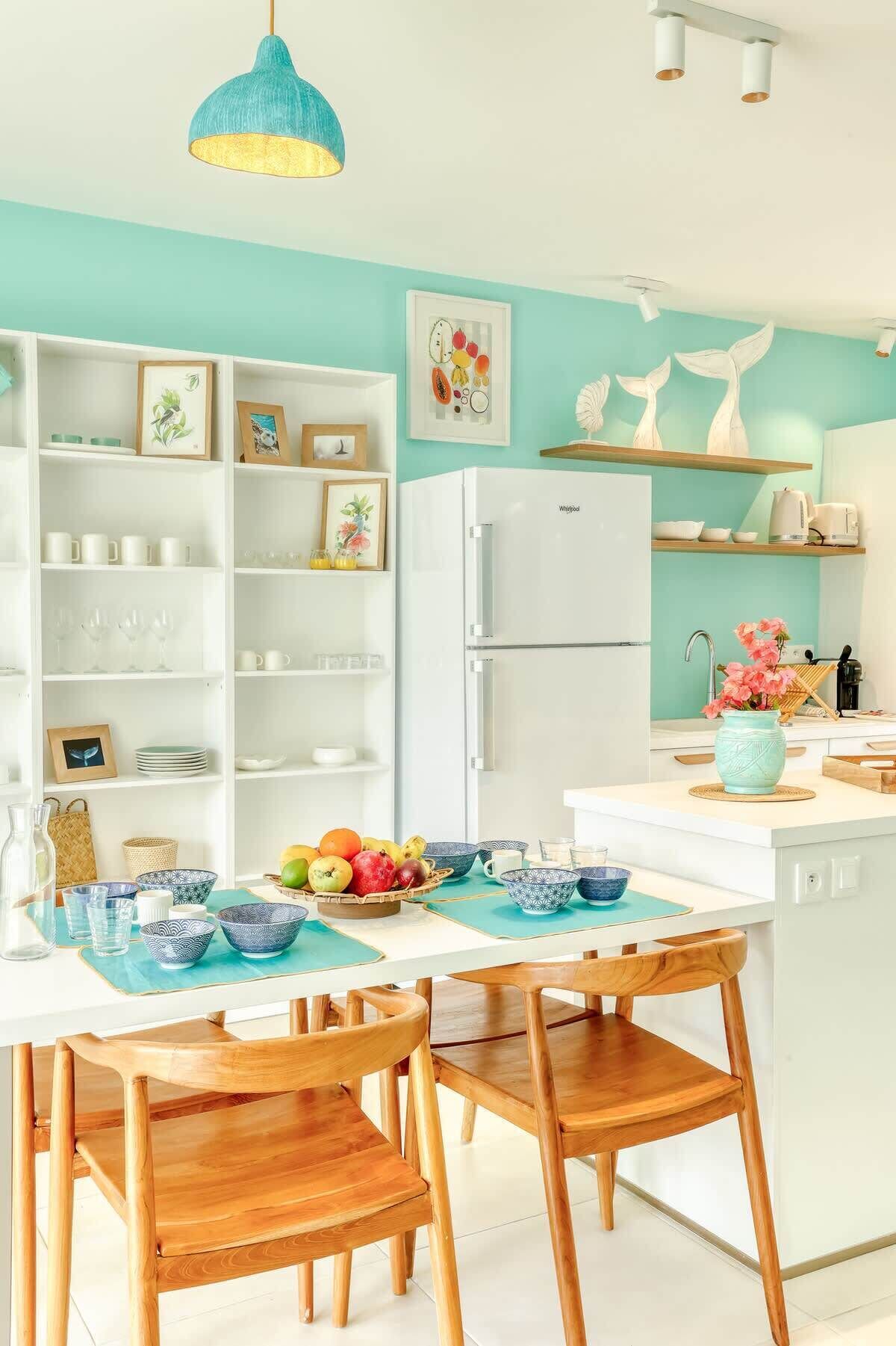 Une cuisine moderne avec des meubles blancs, un réfrigérateur blanc, des murs de couleur turquoise et des chaises en bois.