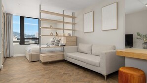 Ein modernes Wohnzimmer mit Fischgrätparkett, einem hellen Sofa, einer Holz-TV-Konsole und einem großen Fenster mit Blick auf die Stadt.