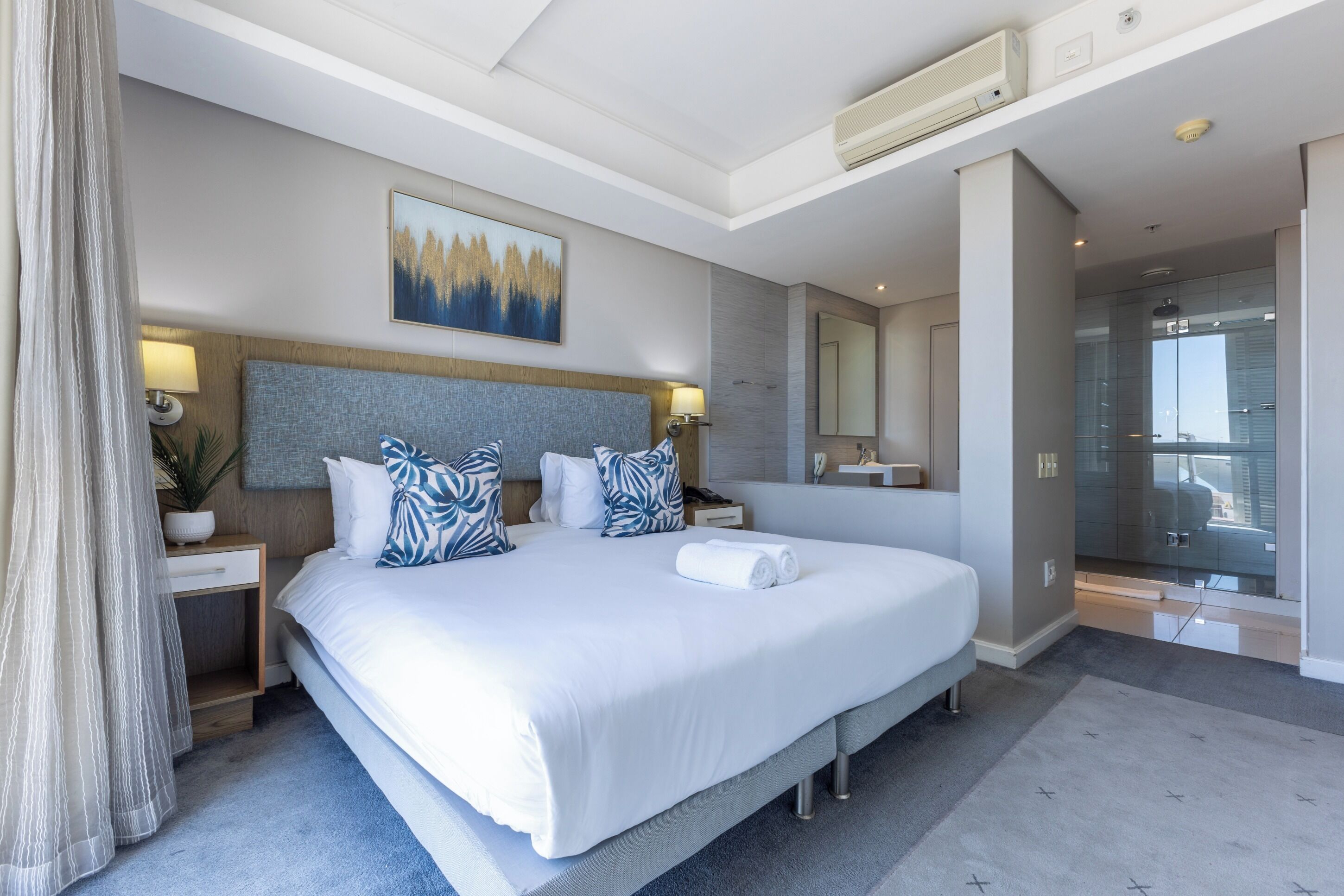 A modern hotel room with a large bed, a bedside table, a nightstand, a wall-mounted TV, and a bathroom visible through an open door.