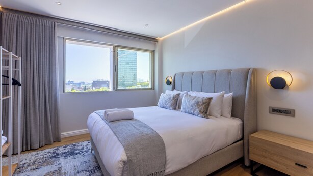 Una camera d'albergo moderna con un grande letto, vista sulla città e un comodino in legno.