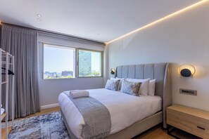 A modern hotel room with a large bed, a view of the city, and a wooden nightstand. - Designer Sea Point Apartment | Pool + Wifi (Cape Town)