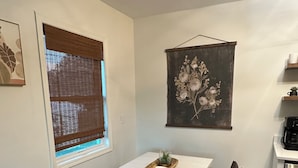 Ett kompakt kök med en matplats, inklusive ett bord med stolar, ett fönster med persienner och en tavla med blommotiv som hänger på väggen.