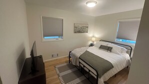 Ett sovrum med en säng, en sänglampa, persienner, en tavla på väggen och en tv på ett tv-bord.
