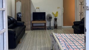 A spacious room with a bed, a sofa, a TV on a stand, a fan, and a door. - Cannarata (Phnom Penh)