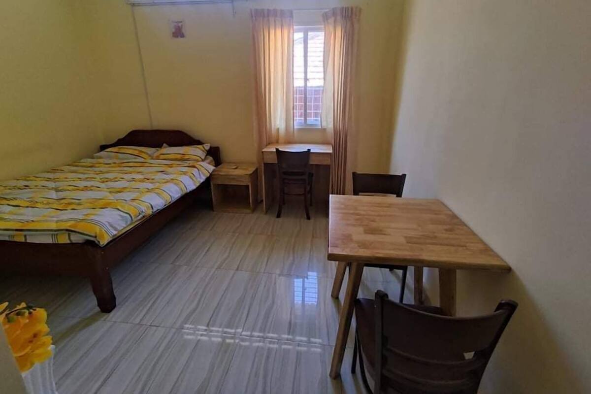Una habitación pequeña con una cama, un escritorio y una silla. Hay una ventana con cortinas y una mesita.