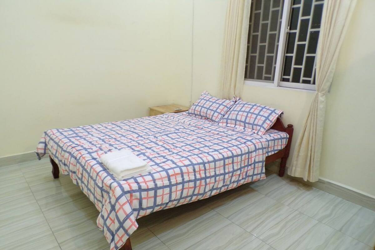 Una cama individual con colcha y almohadas a cuadros, una mesita de noche de madera y una ventana con cortinas.