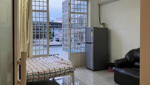 A small, well-lit room with a bed, a refrigerator, a black leather sofa, and a large window with a view of buildings outside. - Cannarata (Phnom Penh)