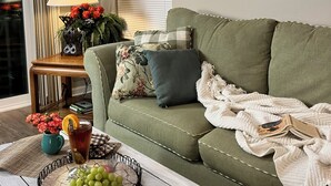 A cozy living room with a green sofa, a side table with a lamp, a framed picture, and a basket of grapes. - Experience the Wild Beauty of Sunset Village from Your Private Balcony (Sunset Beach)