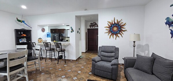 Una sala de estar con un sofá gris, un comedor con mesa y sillas, una cocina con alacenas y un refrigerador, y un adorno de sol en la pared.