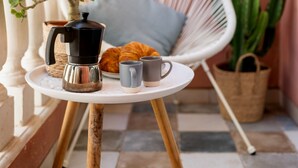 A small round table with a coffee pot, mug, and croissant, a white wicker chair, a cactus, and a macrame wall hanging in a cozy corner.