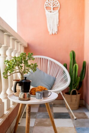 A small round table with a coffee pot, mug, and croissant, a white wicker chair, a cactus, and a macrame wall hanging in a cozy corner.
