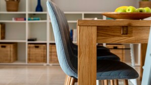 A dining area with a wooden table, a chair with a grey cushion, and a shelf with various items.
