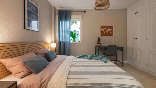 A bedroom with a bed, a desk, a chair, a hanging light, a window with curtains, and a wardrobe.