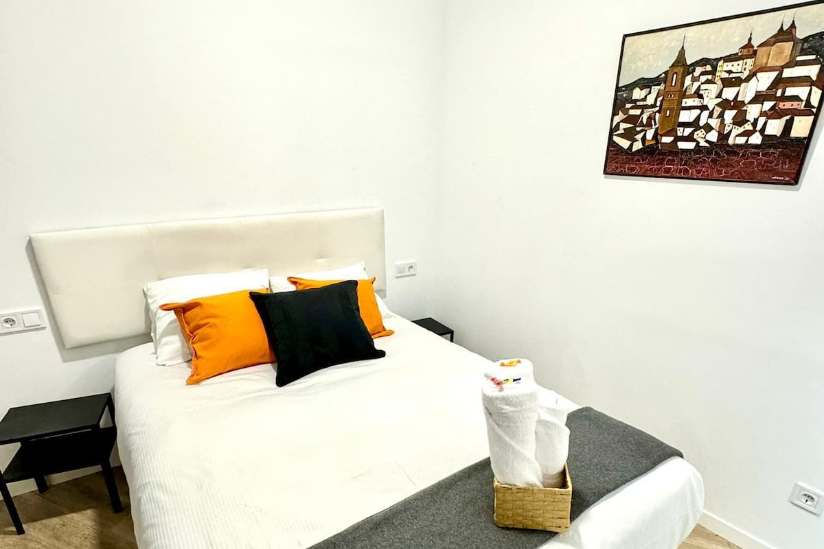 Ein ordentlich gemachtes Bett mit weißer Bettwäsche, einer grauen Decke und orange-schwarzen Kissen. Ein kleiner, schwarzer Nachttisch mit einer Lampe steht neben dem Bett. Ein gerahmtes Kunstwerk eines Gebäudes ist an der Wand.
