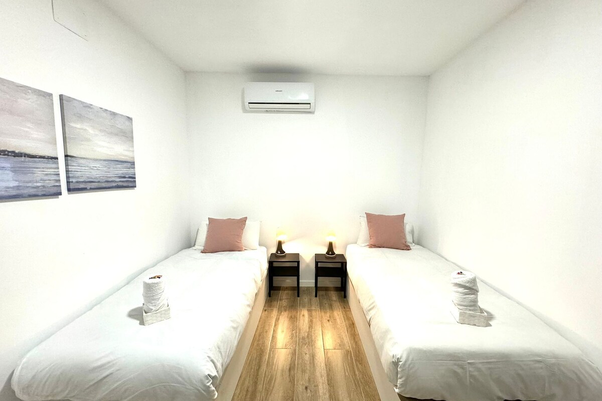 A small, clean room with two single beds, a bedside table with a lamp, and a wall-mounted air conditioner.