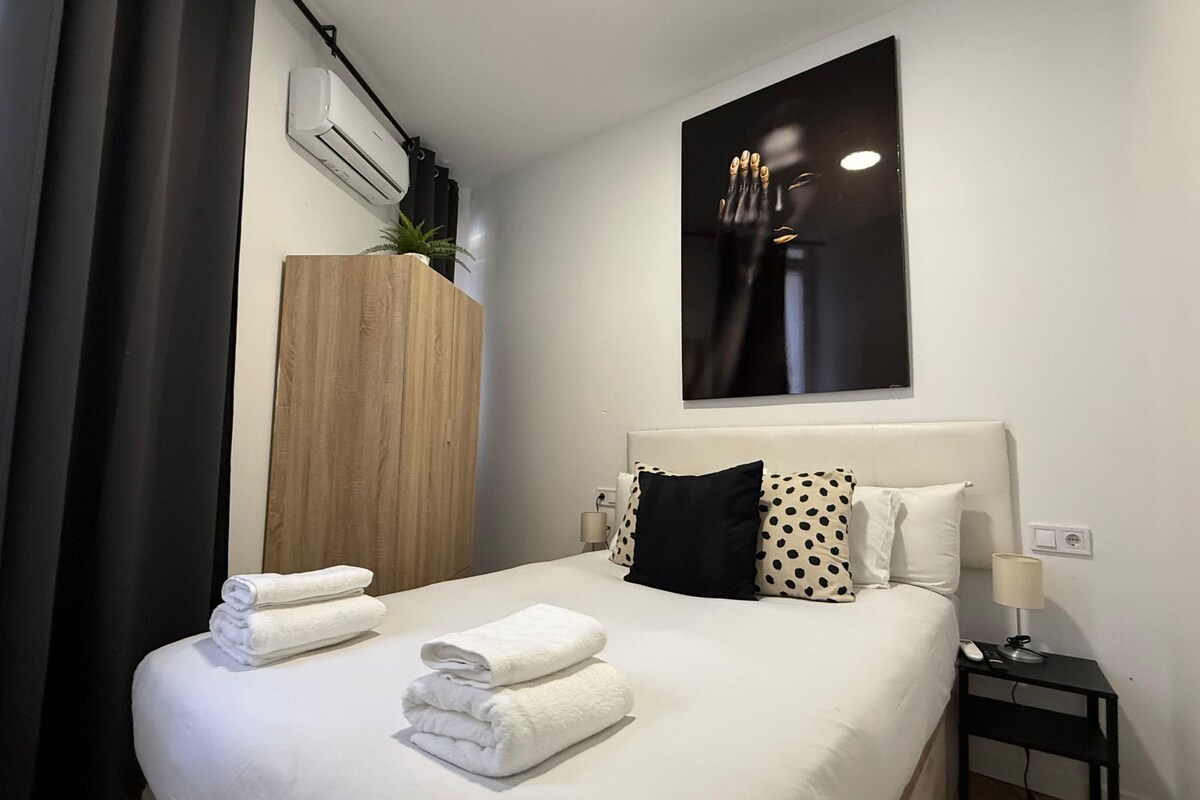 A hotel room with a bed, a nightstand, a lamp, a wall-mounted air conditioner, and a large abstract painting.