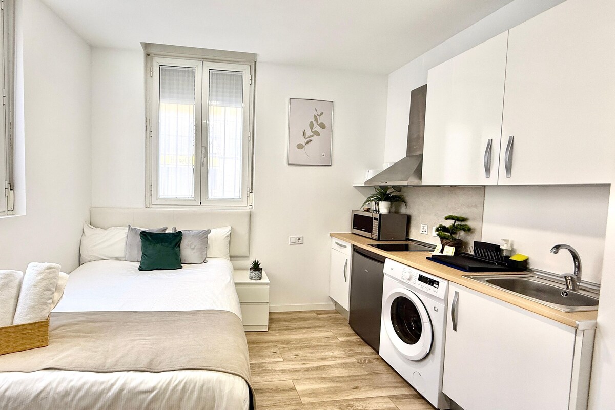 A compact bedroom with a bed, bedside table, and a window. Adjacent is a kitchenette with a washing machine, sink, and microwave.