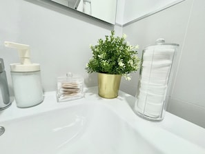 A bathroom sink with a clear glass container, a gold-colored plant, a white bottle, and a clear plastic container with white cotton swabs.
