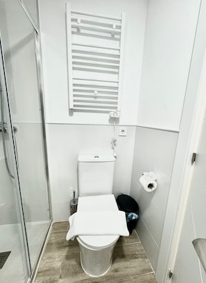 A small, compact bathroom with a shower, toilet, and a window with blinds.