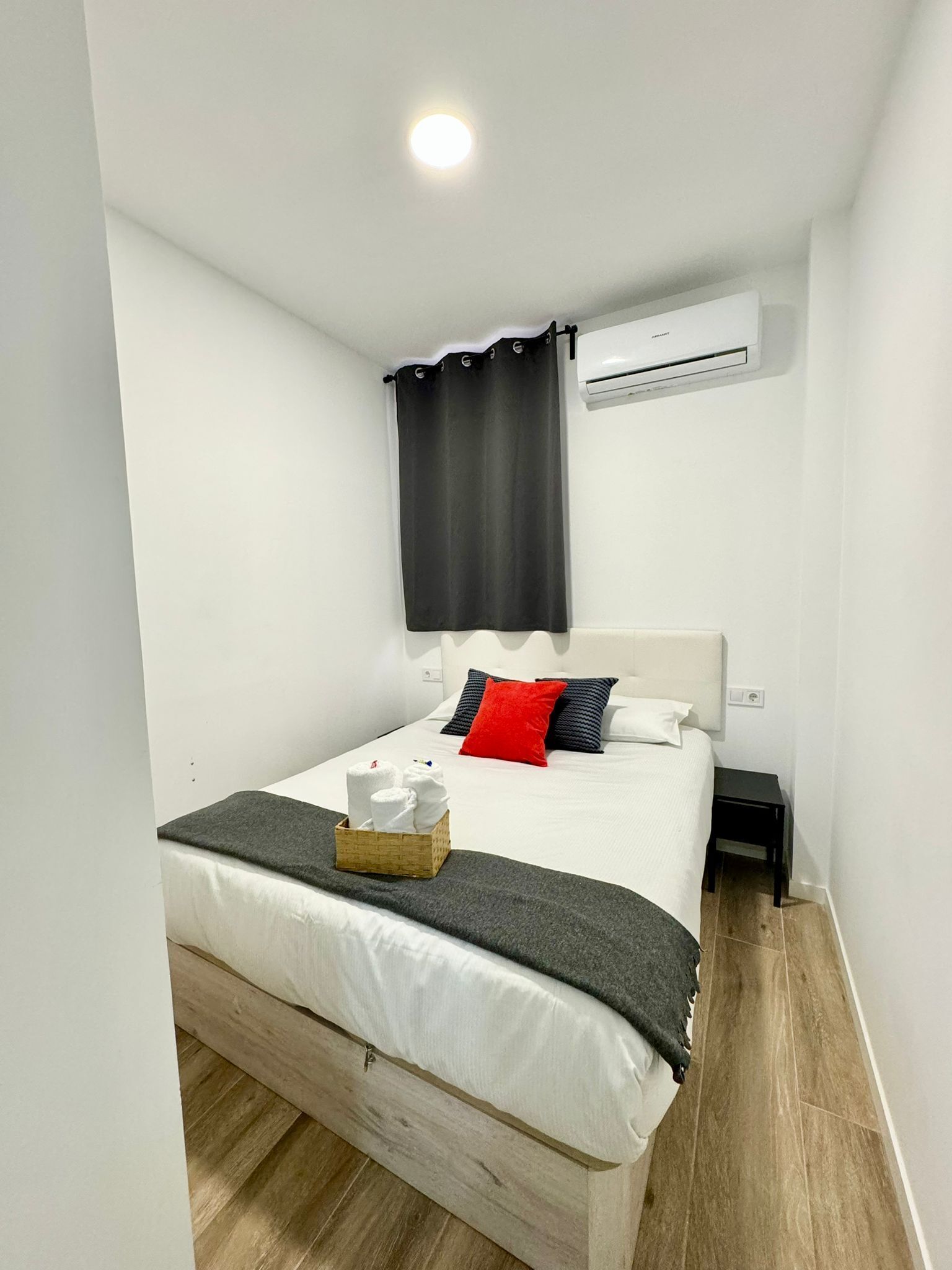 A neatly arranged bedroom with a bed, a bedside table, a basket of towels, and an air conditioning unit on the wall.