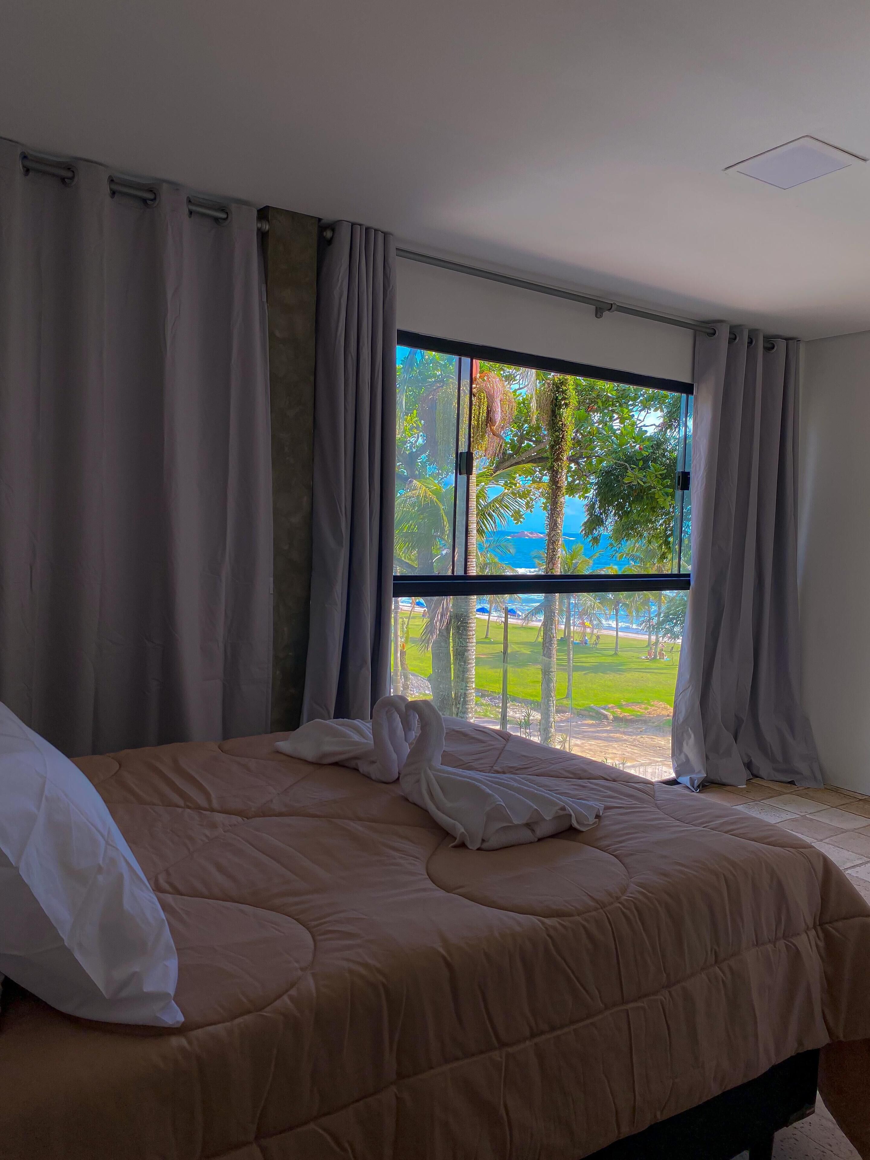 A hotel room with a large bed, a window with curtains, and a view of a beach and palm trees.