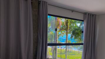 A hotel room with a large bed, a window with curtains, and a view of a beach and palm trees.