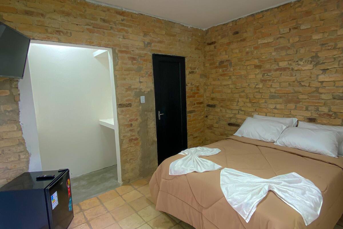 A bedroom with a brick wall, a bed with white linens, a black suitcase, and a door.
