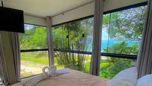 Ett hotellrum med en säng, en tv som är monterad på väggen och ett stort fönster med utsikt över grönområden och en strand.