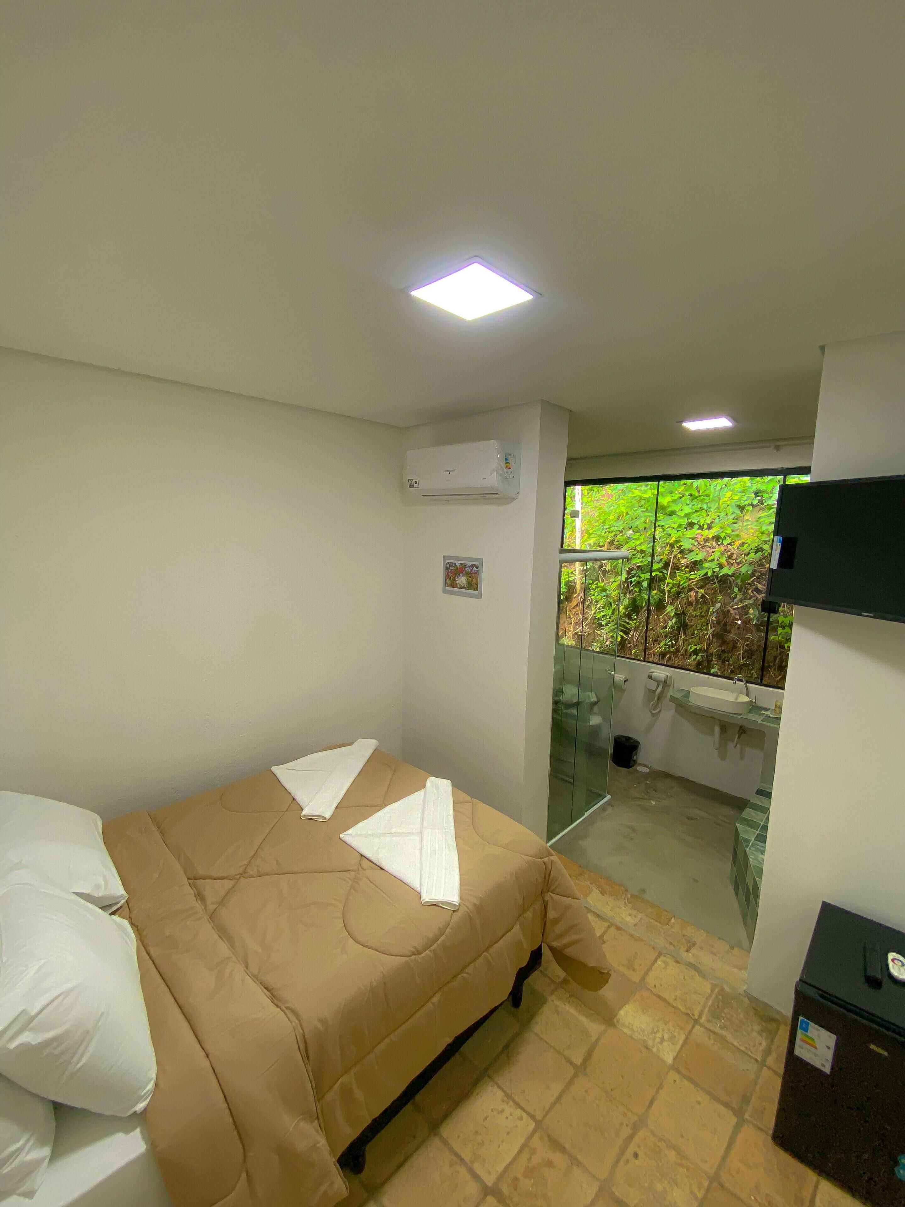 A hotel room with a bed, an air conditioning unit, a small picture on the wall, a sink, and a view of greenery outside.