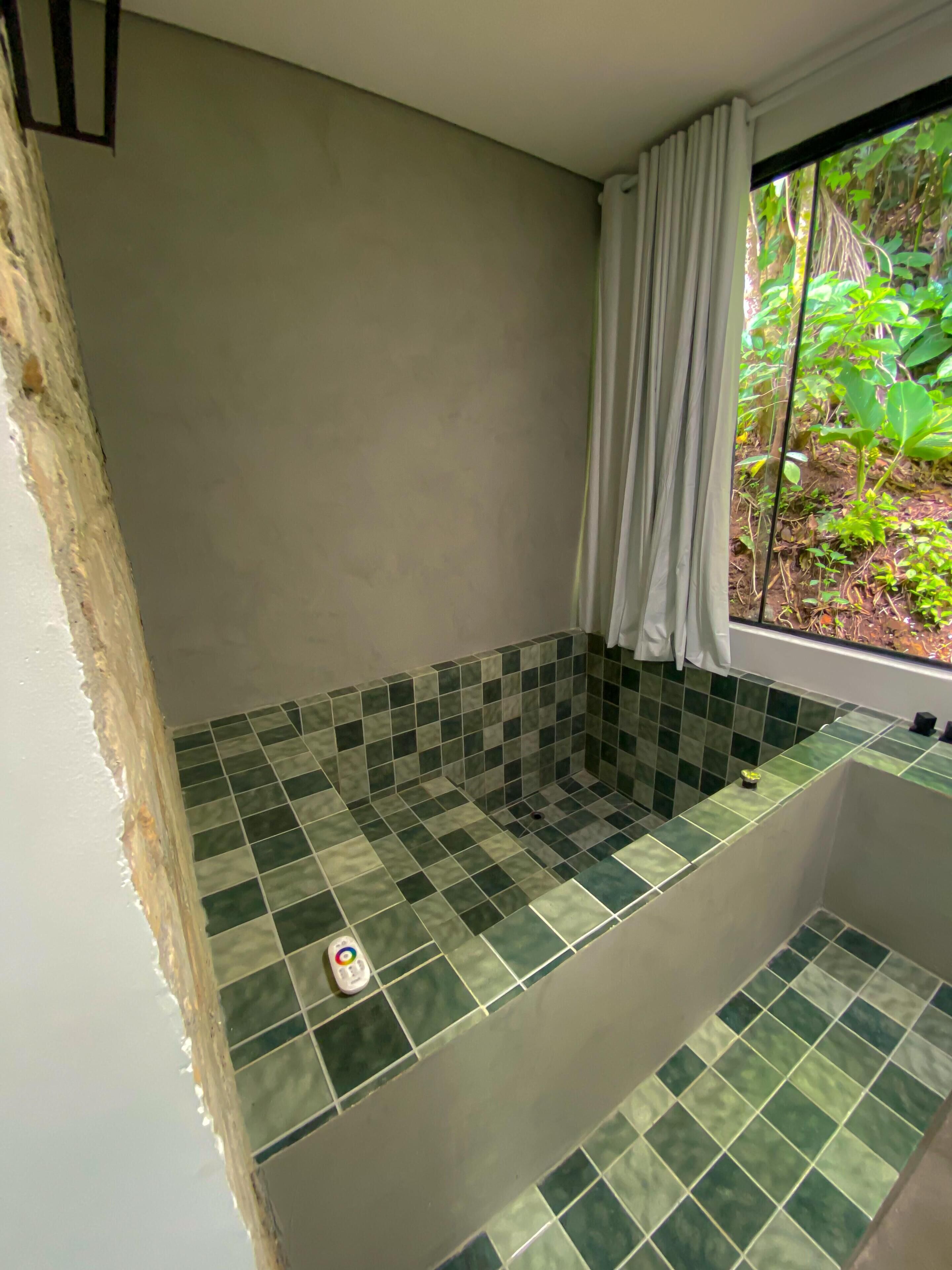 Une salle de bain avec une baignoire recouverte de carreaux verts, une fenêtre avec un rideau blanc et une vue sur la verdure à l’extérieur.