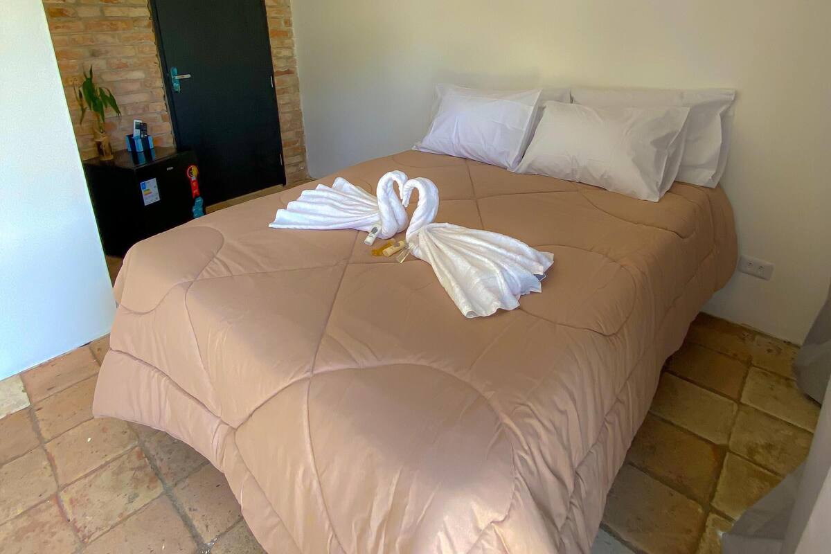 A hotel room with a beige bed, white pillows, and a decorative swan towel arrangement.