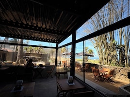 A covered outdoor seating area with tables and chairs, large windows offering a view of the beach and palm trees.