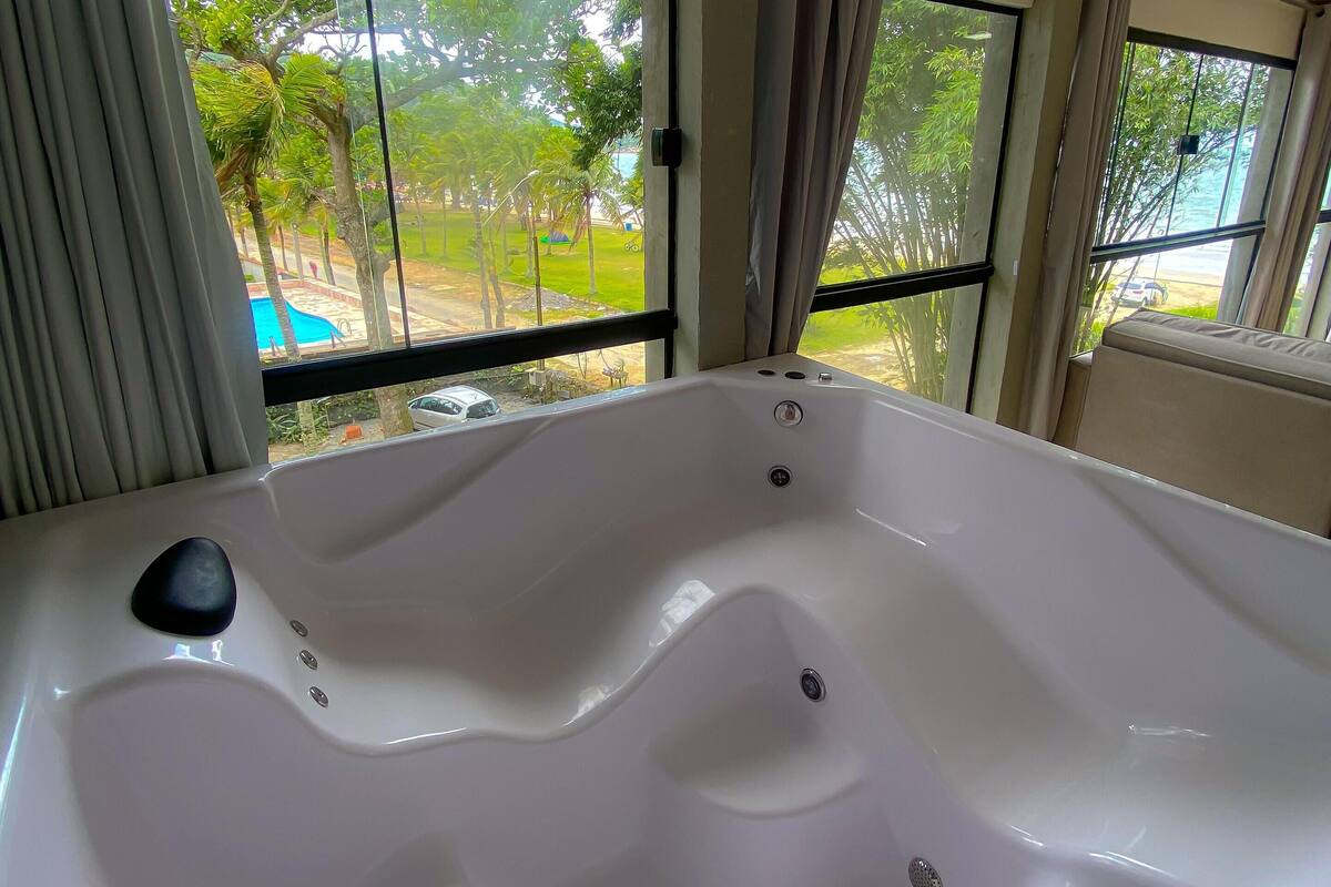 A large white bathtub with multiple jets, positioned near a window with a view of a pool and greenery outside.