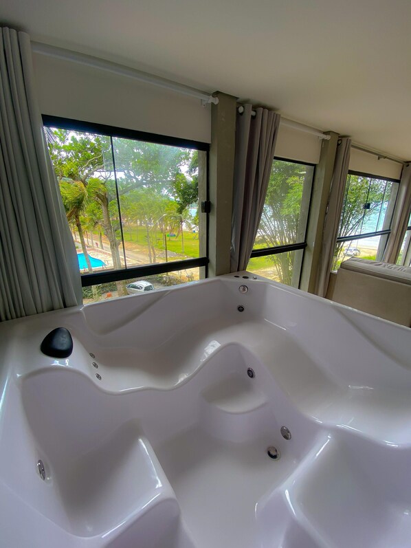 A large white bathtub with multiple jets, positioned near a window with a view of a pool and greenery outside.
