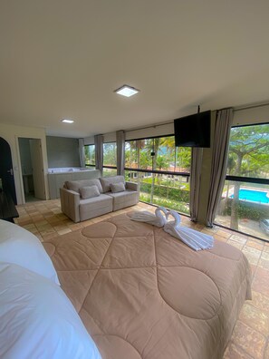 A hotel room with a large bed, a sofa, a TV, and a balcony with a pool view. - Raízes do Mar (Guarujá)