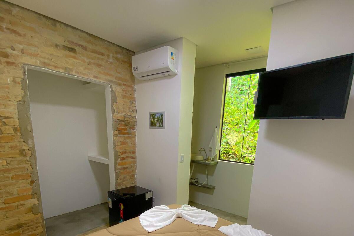 A hotel room with a bed, a TV mounted on the wall, an air conditioning unit, and a window with a view of greenery.