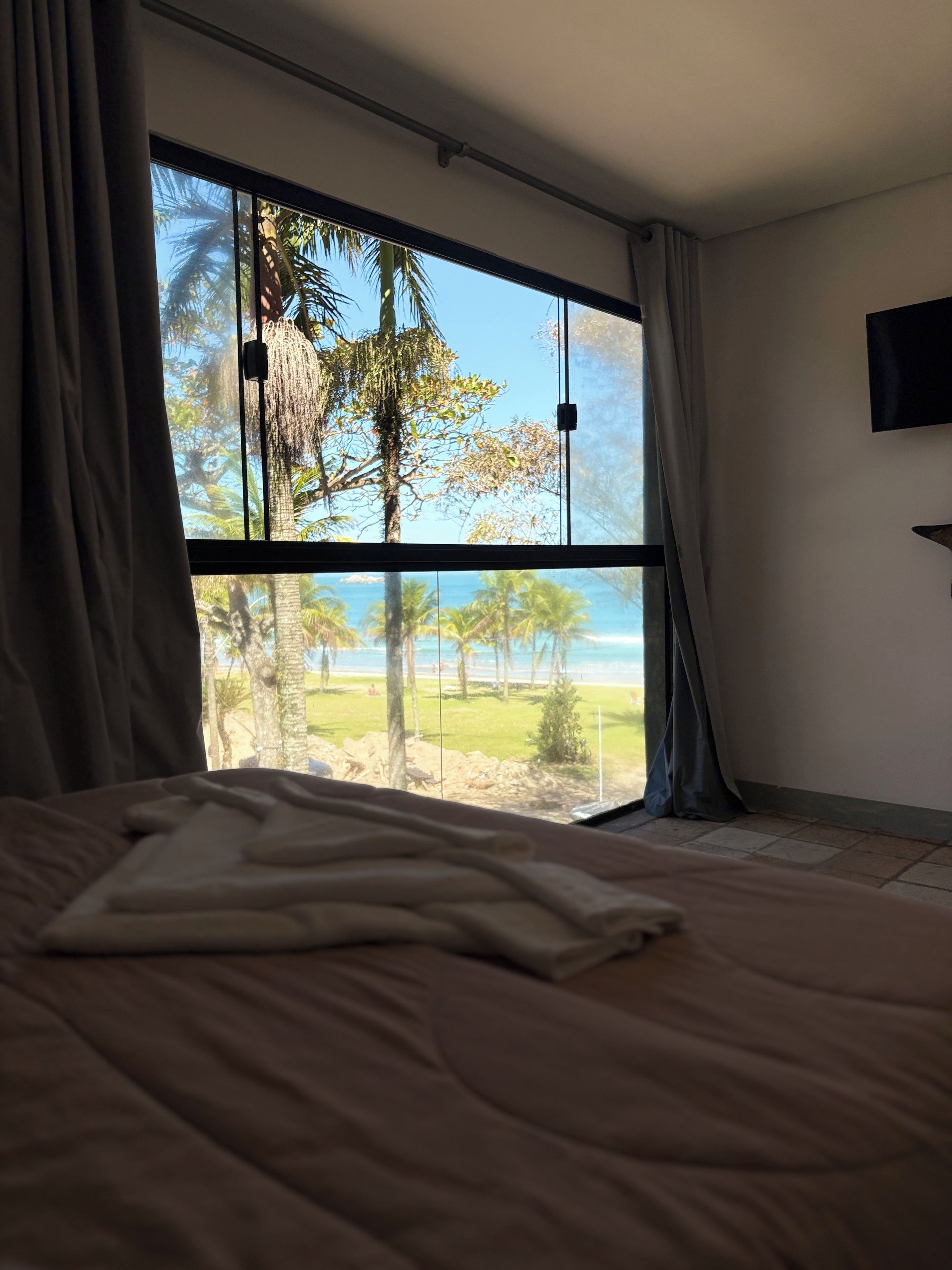 A bedroom with a large window offering a view of the beach and palm trees.