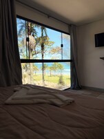 A bedroom with a large window offering a view of the beach and palm trees.