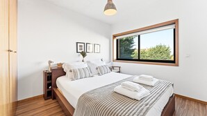 A bedroom with a bed, bedside table, lamp, window, and wall-mounted pictures. - GuestReady - Seaside Experience in Moledo (Moledo)