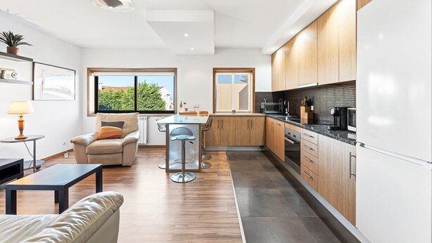 A modern kitchen with wooden cabinets, a dining area with a table and chairs, and a living area with a sofa and a lamp. - GuestReady - Seaside Experience in Moledo (Moledo)