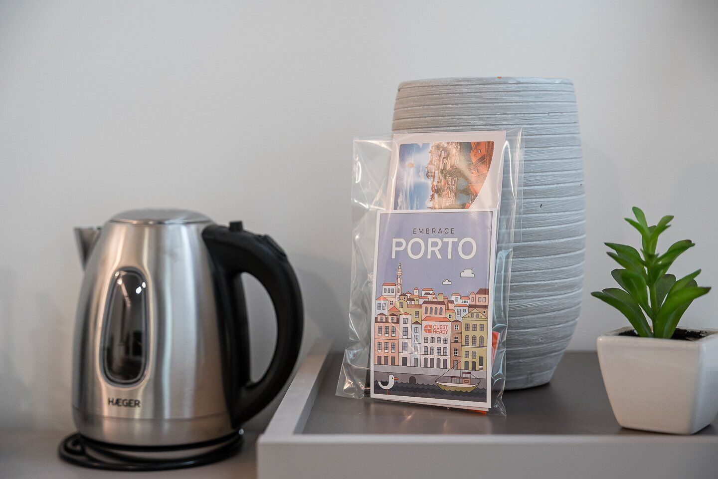 Une bouilloire en acier inoxydable, un guide intitulé « À la découverte de Porto », un grand vase gris et une petite plante en pot sur une table.