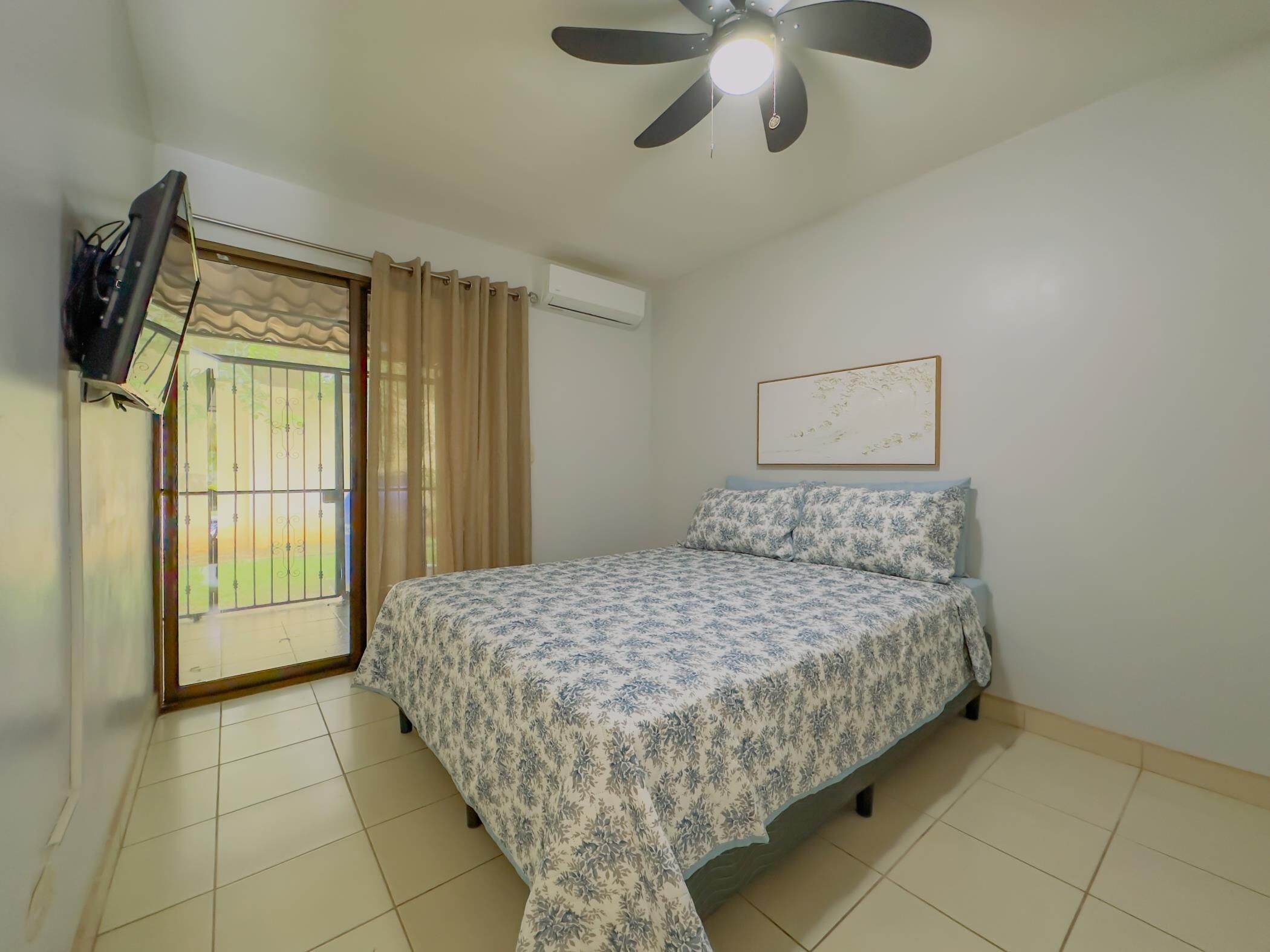 A bedroom with a bed, a ceiling fan, a TV mounted on the wall, and a window with curtains.