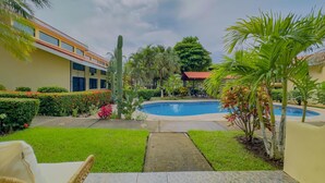 A pool area with a clear view of the pool, surrounded by well-maintained greenery and a building with a red-tiled roof.