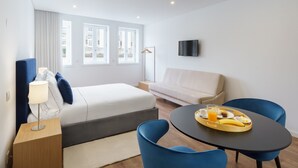 A modern hotel room with a bed, a bedside table with a lamp, a round table with breakfast items, a sofa, and a TV on the wall. - Feel Porto Lapa Collection Flat 2 2 (Porto)