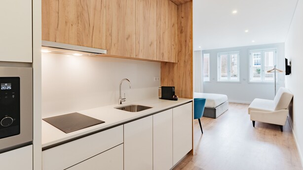 A modern kitchen with white cabinets, a wooden upper section, and a built-in oven. There is a sink with a faucet, a countertop, and a coffee machine. The room has a large window, a white sofa, and a blue chair. - Feel Porto Lapa Collection Flat 1 2 (Porto)